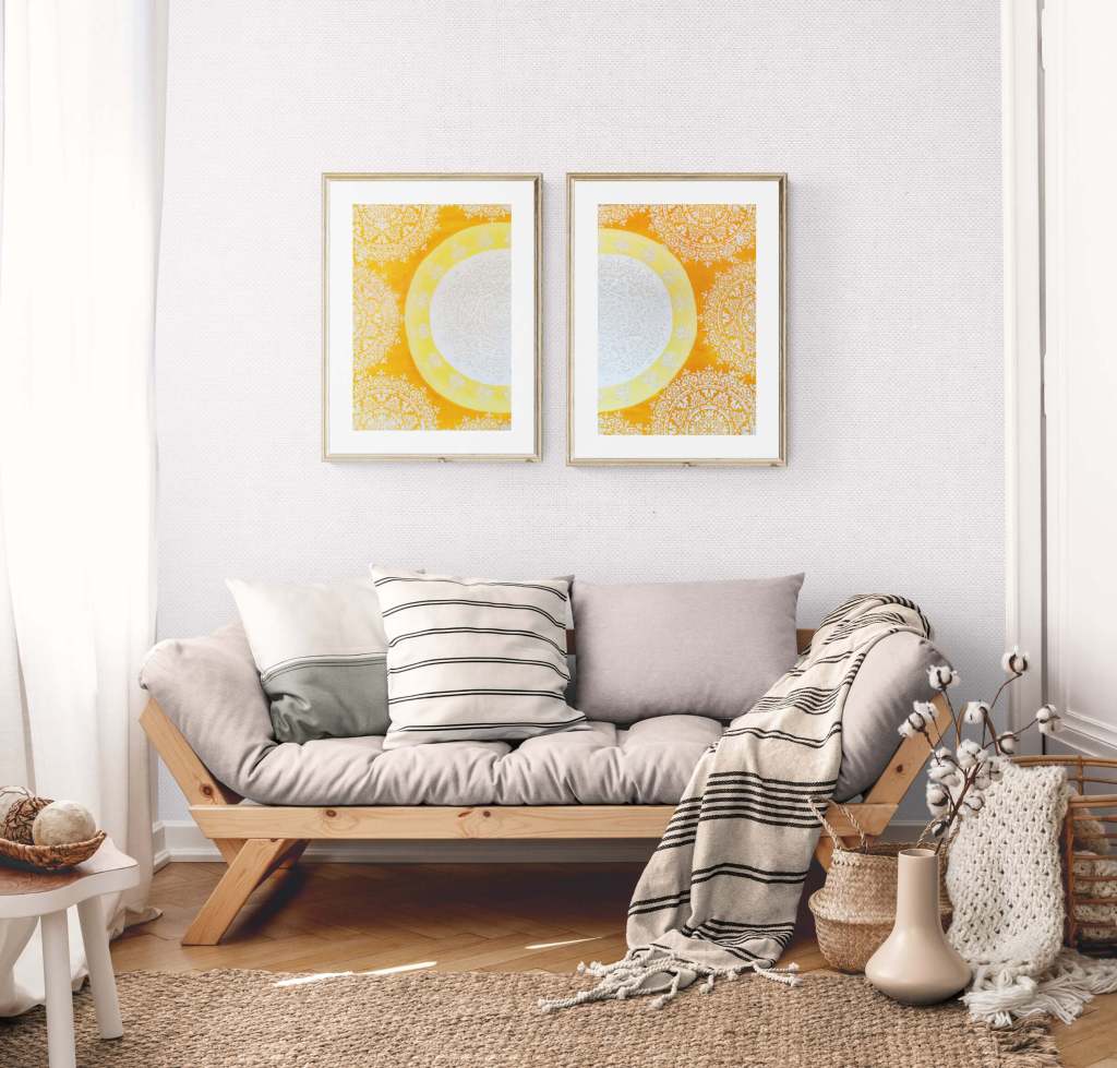 Showcasing two paintings on top of a sofa, featuring A Sun Mandala divided in two, in white, yellow, and orange with sparkles.