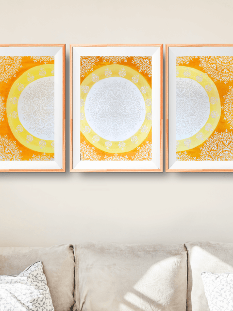 Showcasing a triptych on top of a sofa, featuring A Sun Mandala divided in three, in white, yellow, and orange with sparkles.
