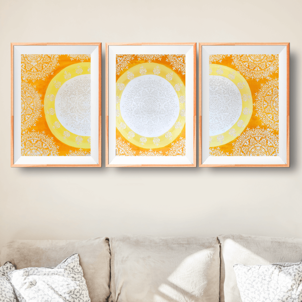 Showcasing a triptych on top of a sofa, featuring A Sun Mandala divided in three, in white, yellow, and orange with sparkles.